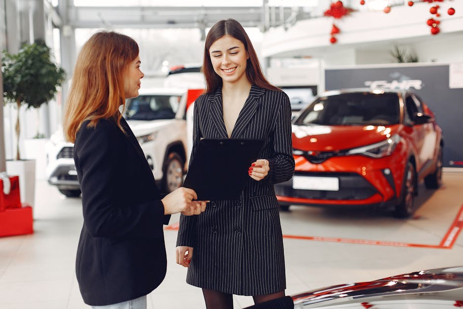 Cheerful young friendly dealer in formal stylish black dress showing contract to smiling female customer in black jacket while standing in car showroom again…